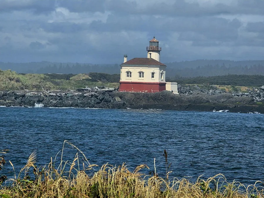 The Coquille River Lighthouse on a stormy afternoon, the white tower and red base standing on a basalt jetty with driftwood piled along the shore
